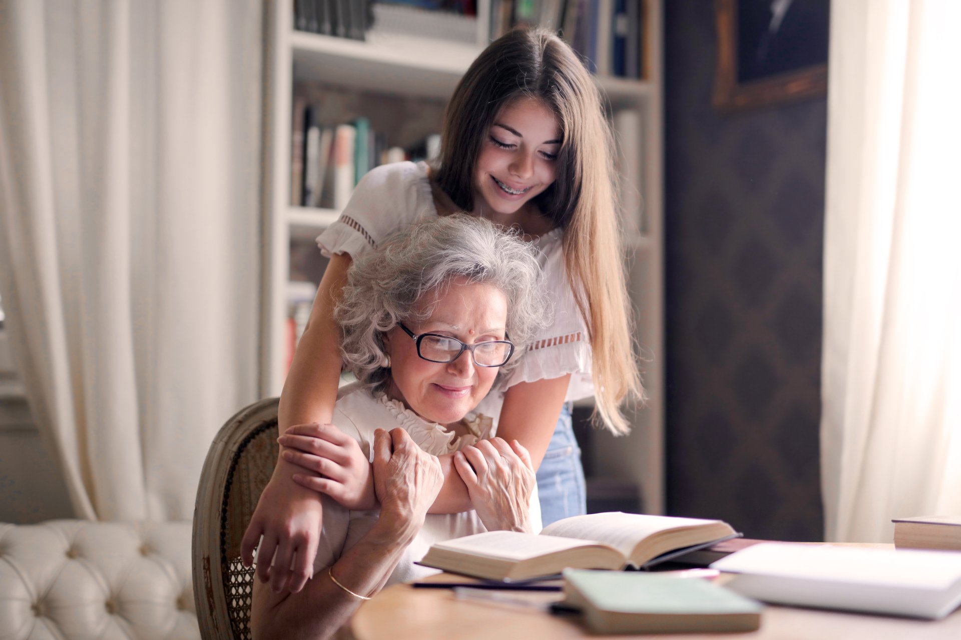A young woman hugging an older woman who is reading at a table