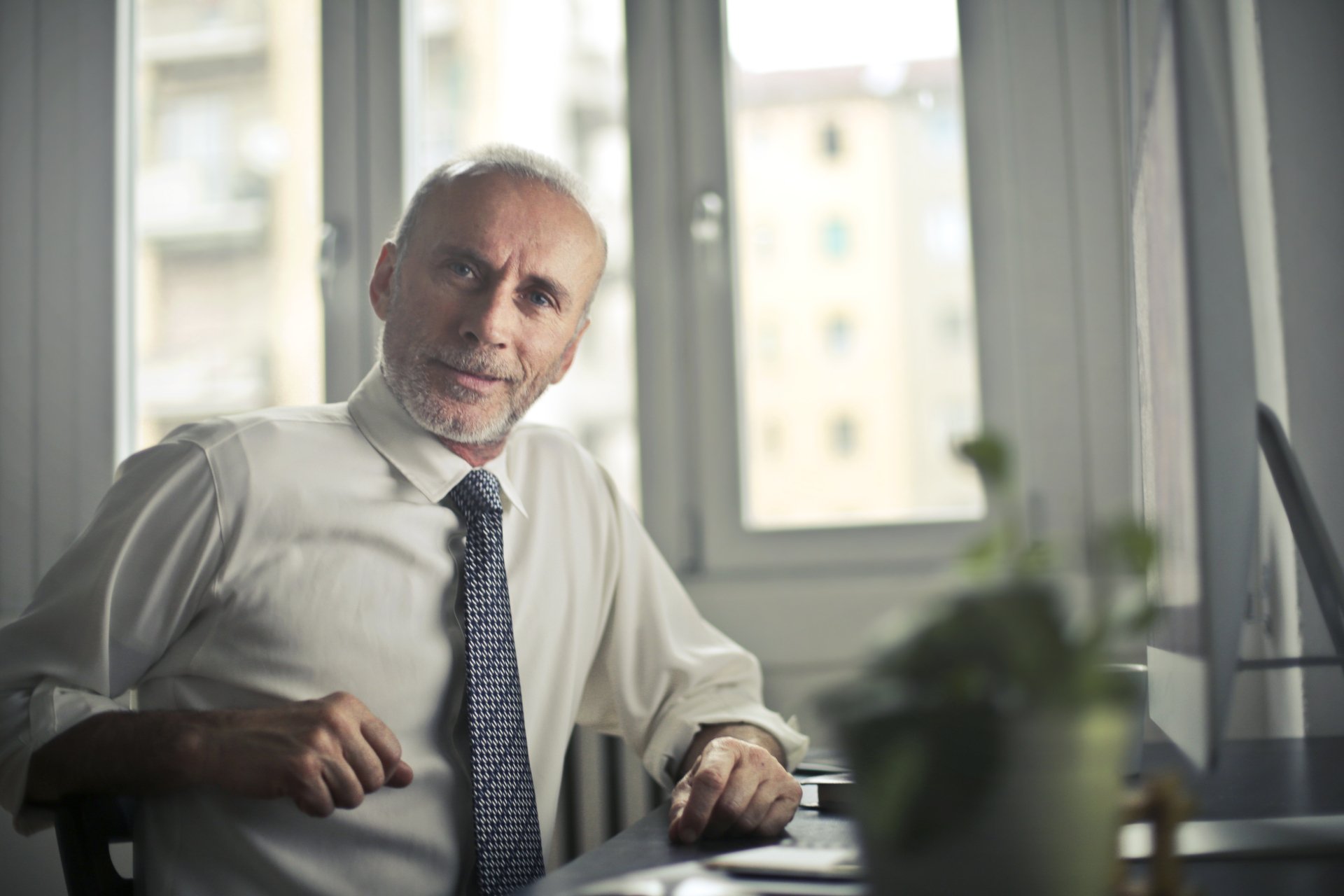 A smiling man in a shirt and tie sitting at a desk by a window