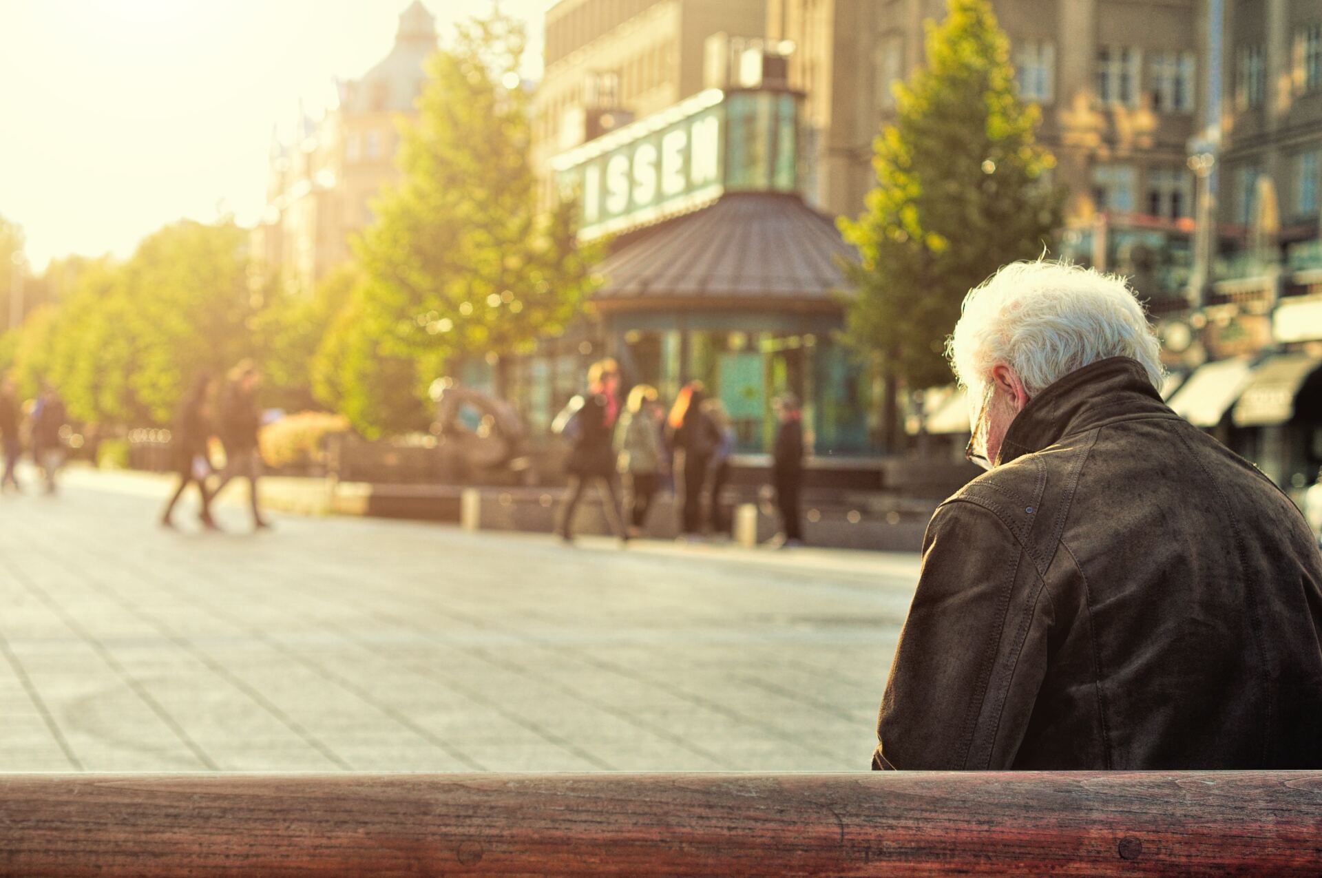 An older man in a jacket sitting on a bench looking at a city square